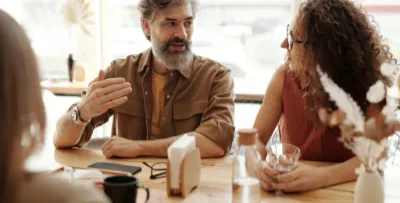 Eine Gruppe Menschen verschiedenen Alters sitzt an einem Tisch. Im Fokus stehen ein älterer, weißhaariger, bärtiger Mann in orangem T-Shirt mit braunem, offenen Hemd darüber und eine junge Frau in einem roten Oberteil mit lockigen Haaren. Die beiden kommunizieren angeregt.