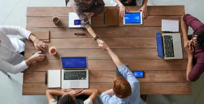 Vogelperspektive eines Holztisches, auf dem mehrere Menschen an Laptops arbeiten. Zu sehen sind nur die Hände und Unterarme der Personen sowie die Laptops und einige Notizbücher. Zwei der Personen reichen sich über die Mitte des Tisches hinweg die Hand und schütteln sie.