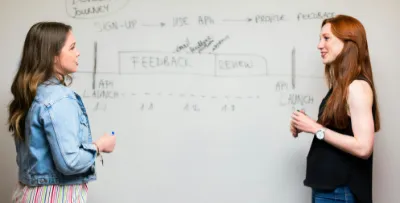 Zwei junge Frauen stehen vor einem Whiteboard. Auf dem Board sind mehrere Scribbles verschwommen erkennbar. Die beiden Frauen tauschen sich angeregt aus.